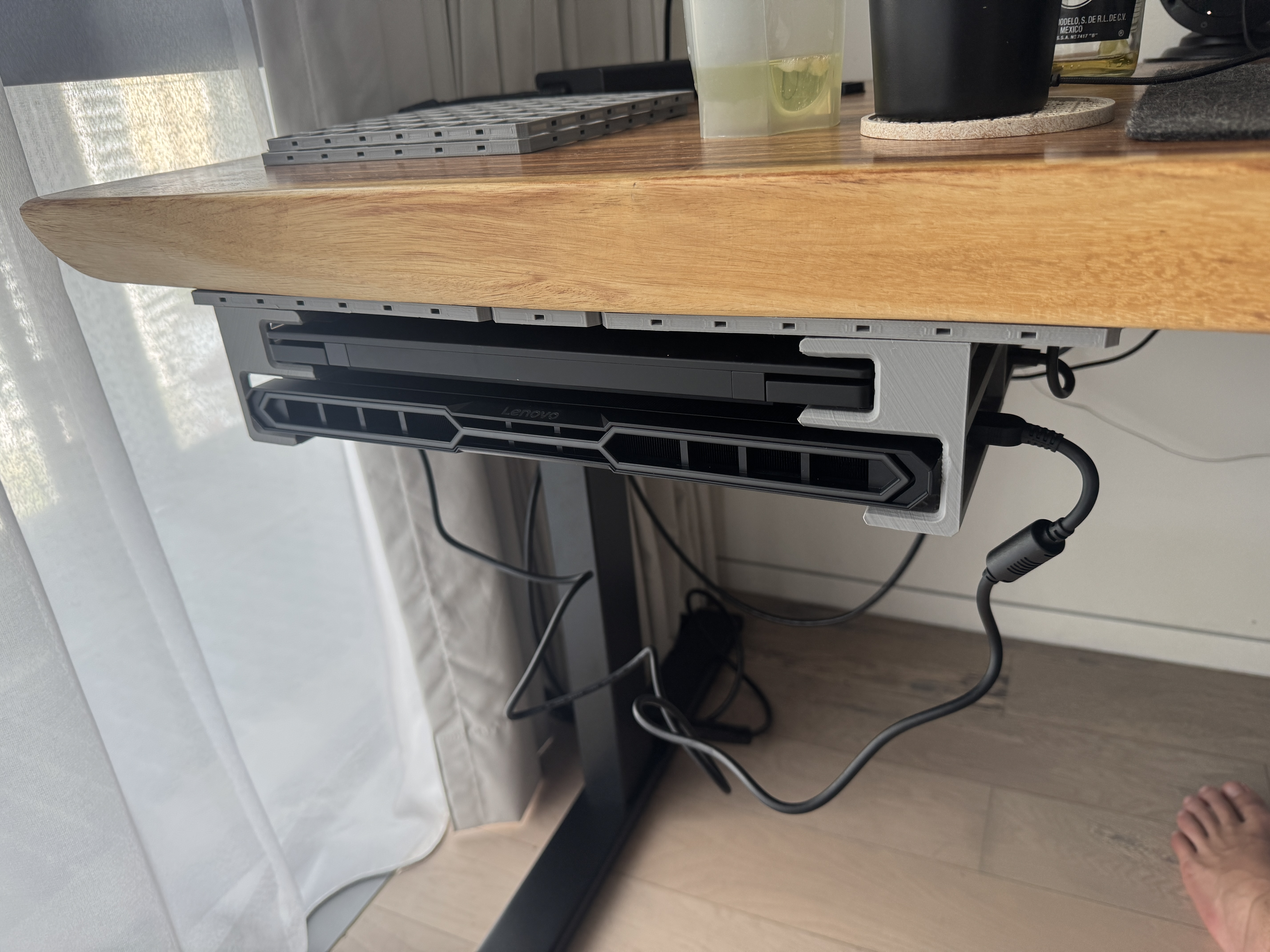 A grey 3D-printed openGrid tray fixed beneath a wooden desk, holding a black Lenovo laptop with its charger plugged in.